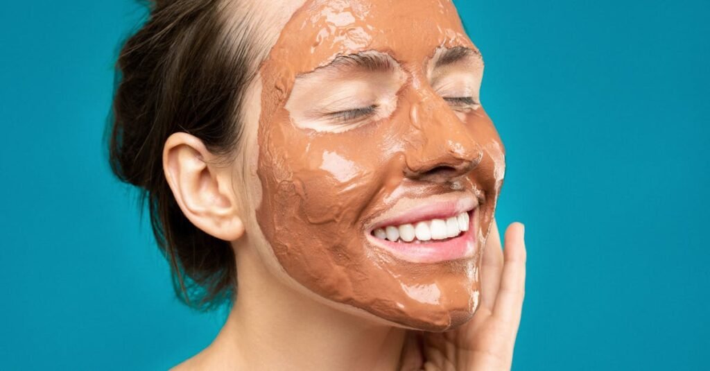 Close-up of a woman smiling with a clay mud mask on her face on a turquoise background.